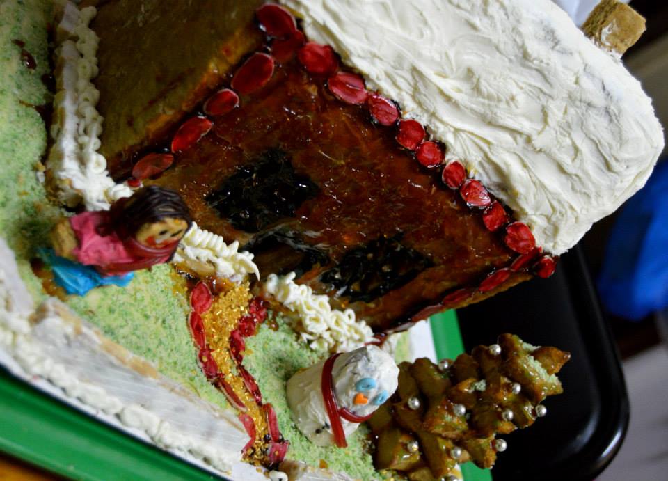 A crooked picture of a homemade gingerbread house. The roof is of a white iceing badly spread, the walls are uneavenly covered in caramel, there is a gingerbread man with a face so badly drawn is almost scary and a white truffe snowman that looks quite sad.