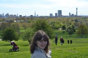 A portrait of Iris (the author) on a green hill. She is a brunette with straight sleek bob, fair skin and big smile. There are some couples walking behind her. London skyline is in the background.