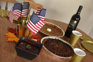 A wooden table featuring a black bowl with cranberry sauce, a pecan pie, a pumkin pie with marshmallows, and a bottle of red wine. The table is set up with golden cardboard and plastic dinnerware, and decorated with American flags and fake maple leaves in orange, yellow and red tones.