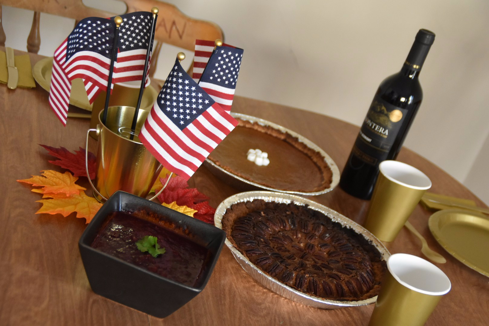 A wooden table featuring a black bowl with cranberry sauce, a pecan pie, a pumkin pie with marshmallows, and a bottle of red wine. The table is set up with golden cardboard and plastic dinnerware, and decorated with American flags and fake maple leaves in orange, yellow and red tones.