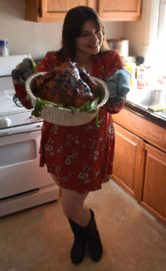 Iris Permuy holding the tray with the turkey ready to serve. She is smiling, she is wearing a floral orange dress, cowboy boots and teal oven gloves.