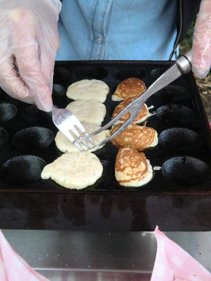 A cast iron pan with four rows of five half-spherical moulds. In the second row from the left there are four mini pancakes still uncooked, which a person with plastic gloves is turning around with a dessert fork and a deli fork. On the next row, there are other four mini pancakes already golden.