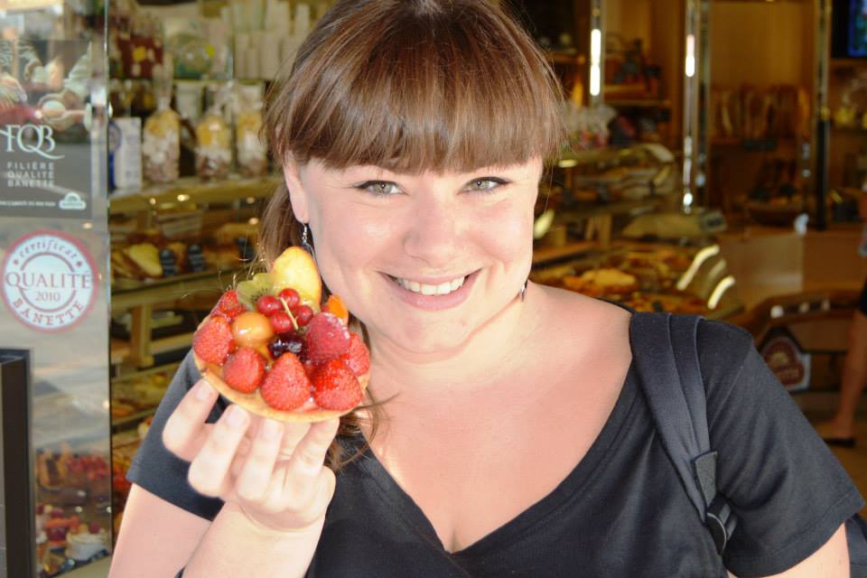 Iris, with a big smile, holding a small tart with strawberries, kiwi, peach, apple, a cherry, a raspberry and some bilberries on it.