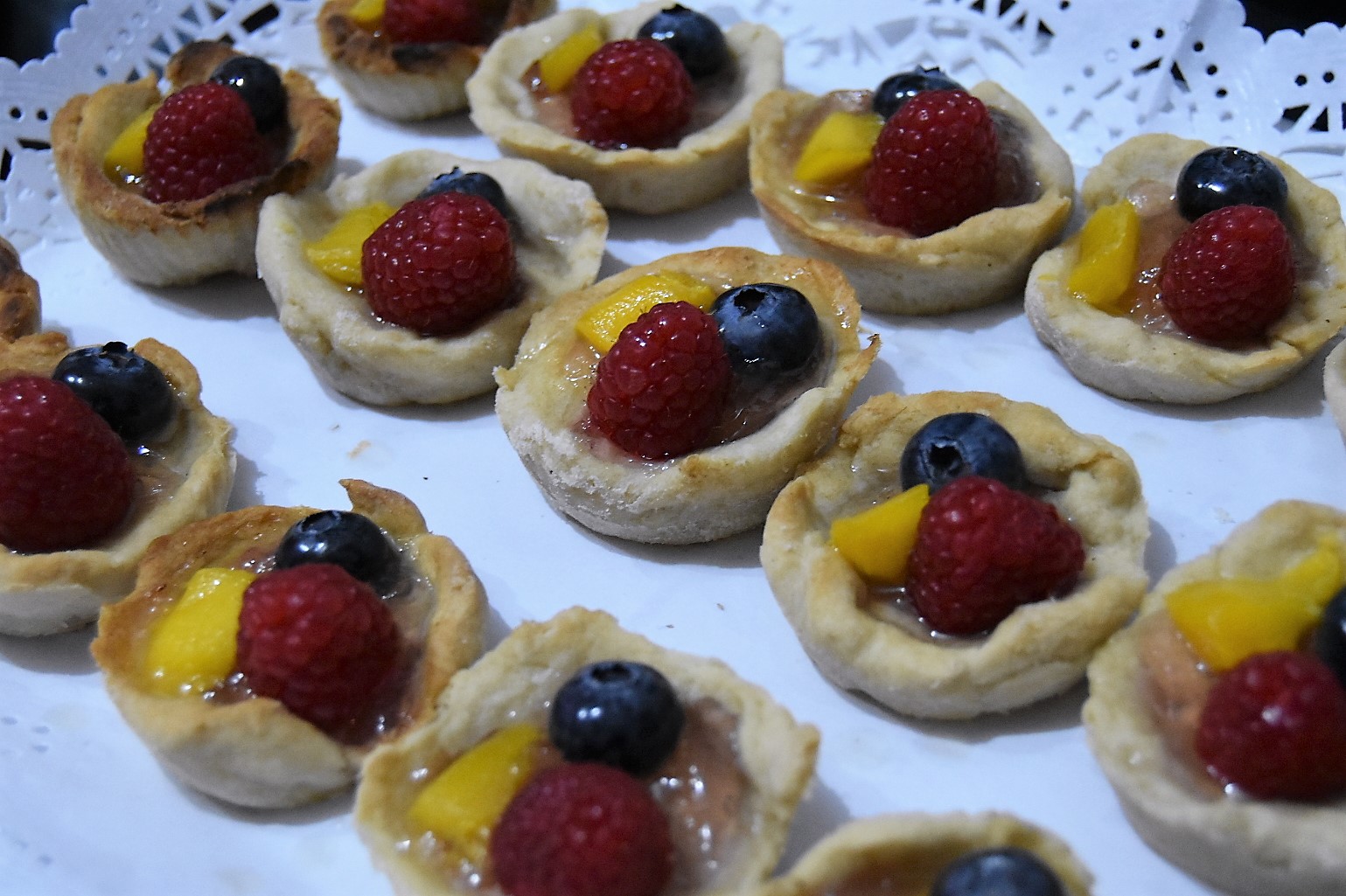 A white plate with three rows of small homemade tarts, each topped with a rasberry, a bluebery and a small piece of mango.