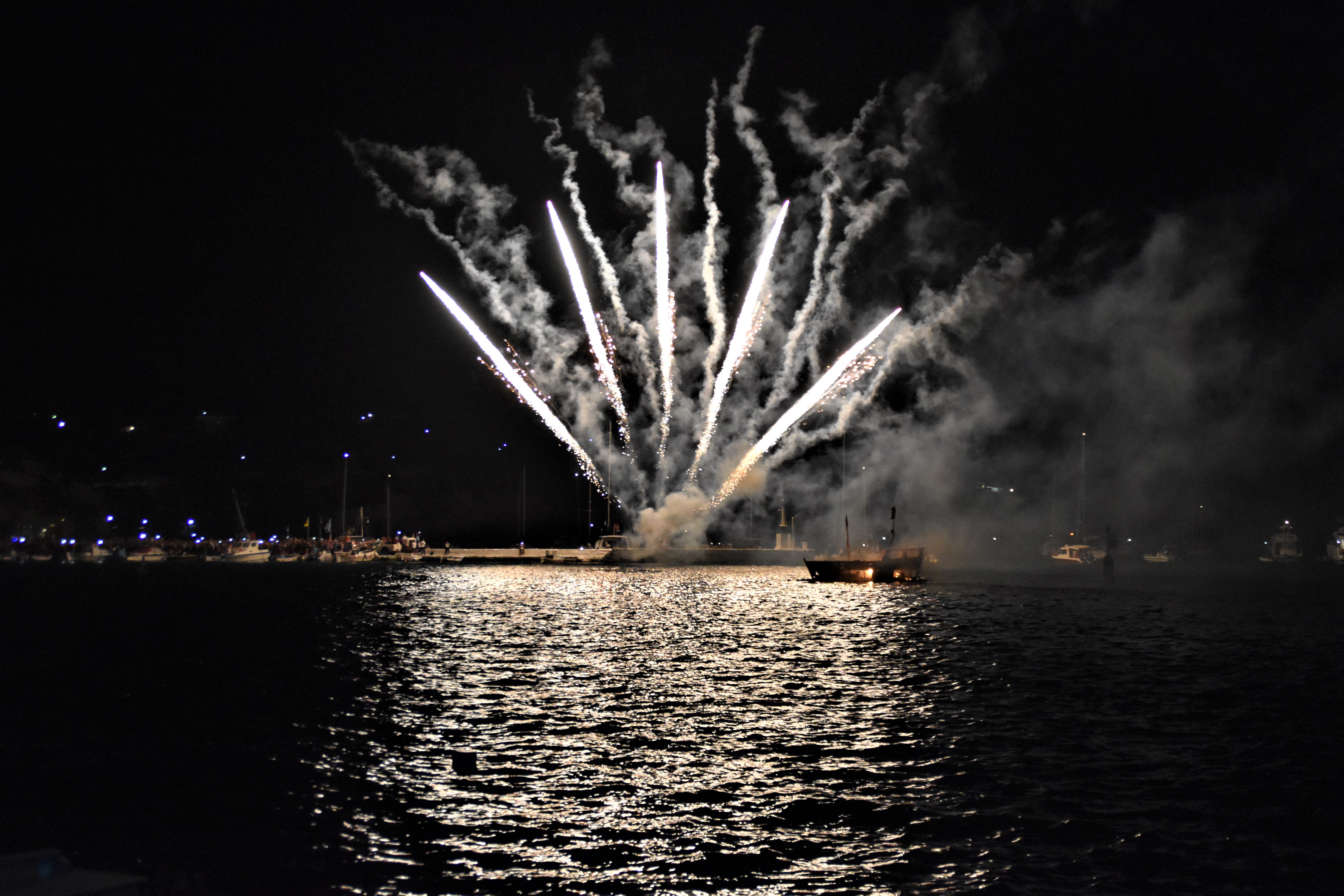 White fireworks emerging from a dark sea illuminate a small burnt boat with glowing embers.