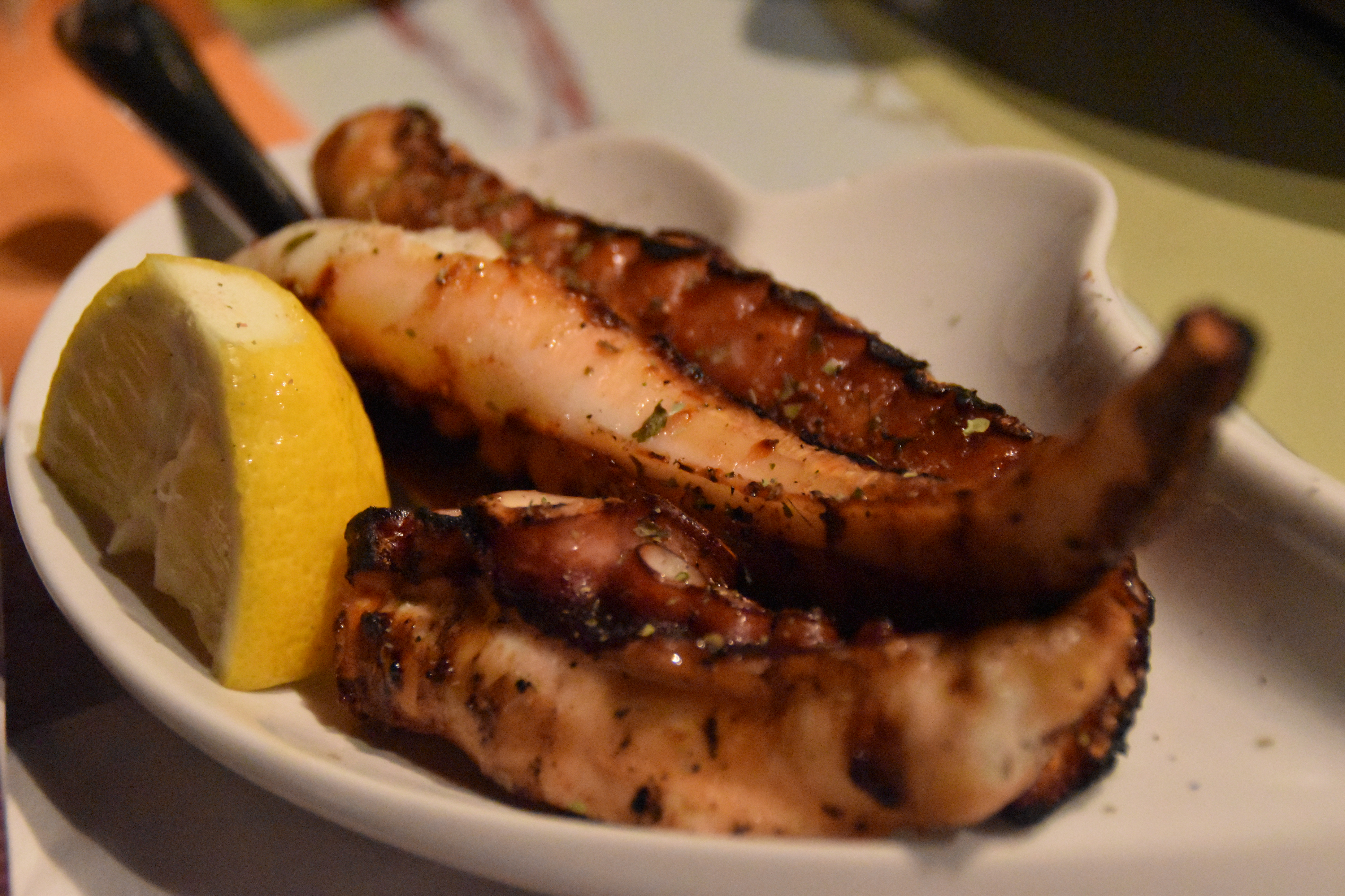 Three thick reddish octopus legs, sprinkled with hebs, served in a white plate with a quarter of a lemon on the side.