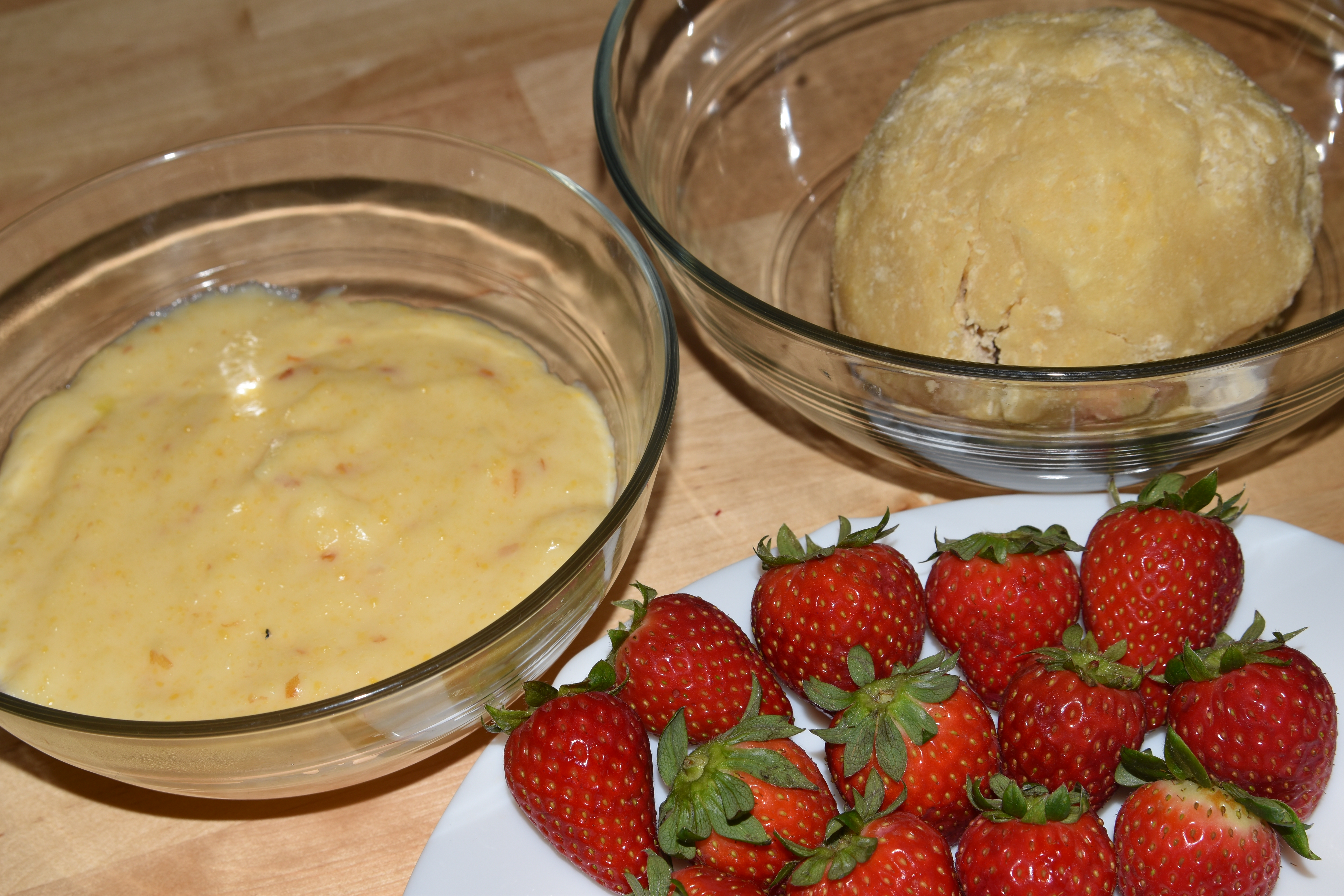 In two separate glass bowls, there is some custard and a ball of pie dough. Next to them, to the right, there is a white plate with strawberries.
