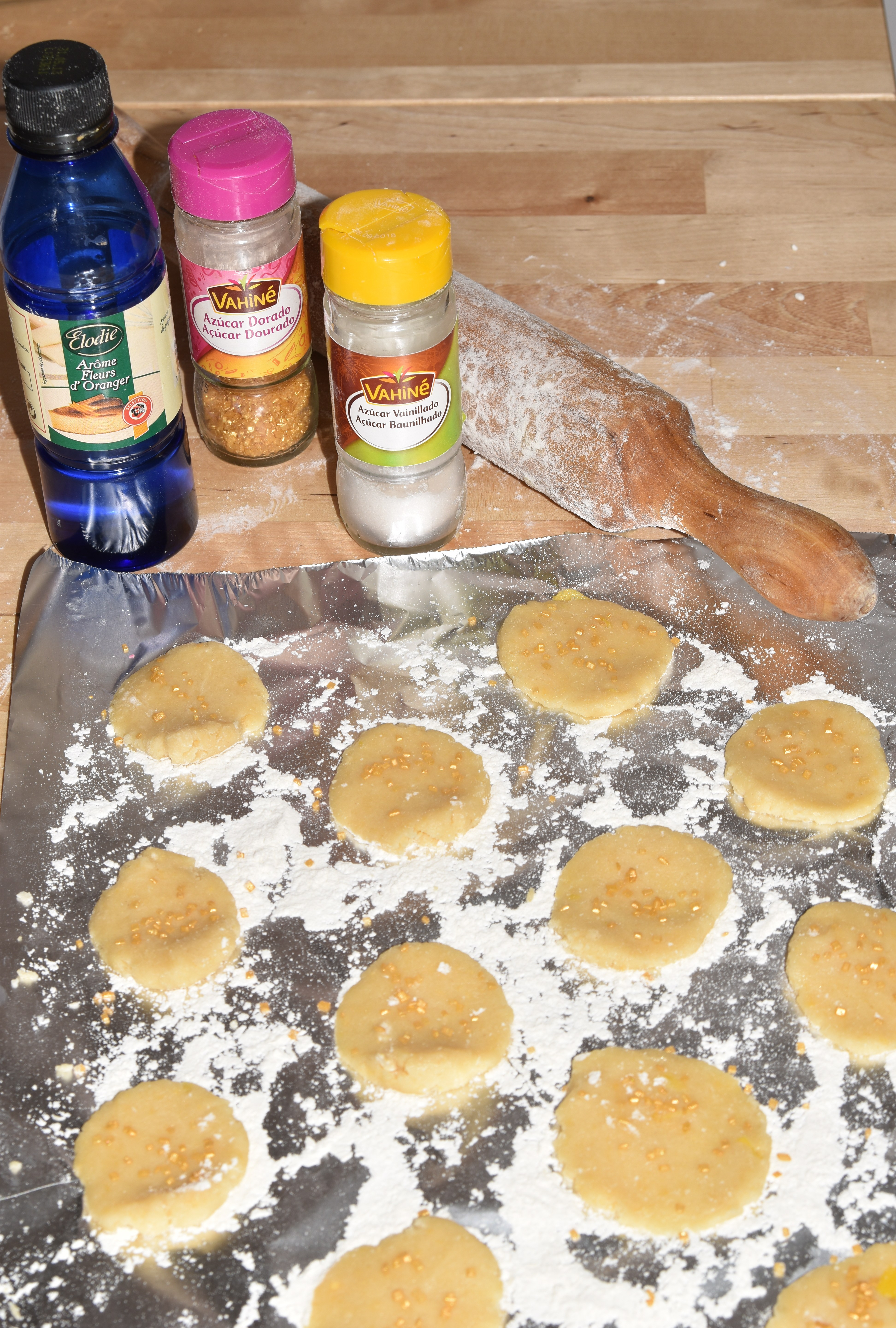 Unbaked cookies laying on a aluminium sheet with flour. Next to it, a plastic bottle of orange blossom water, a golden sugar jar, and a vanilla sugar jar stand next to the rolling pin.