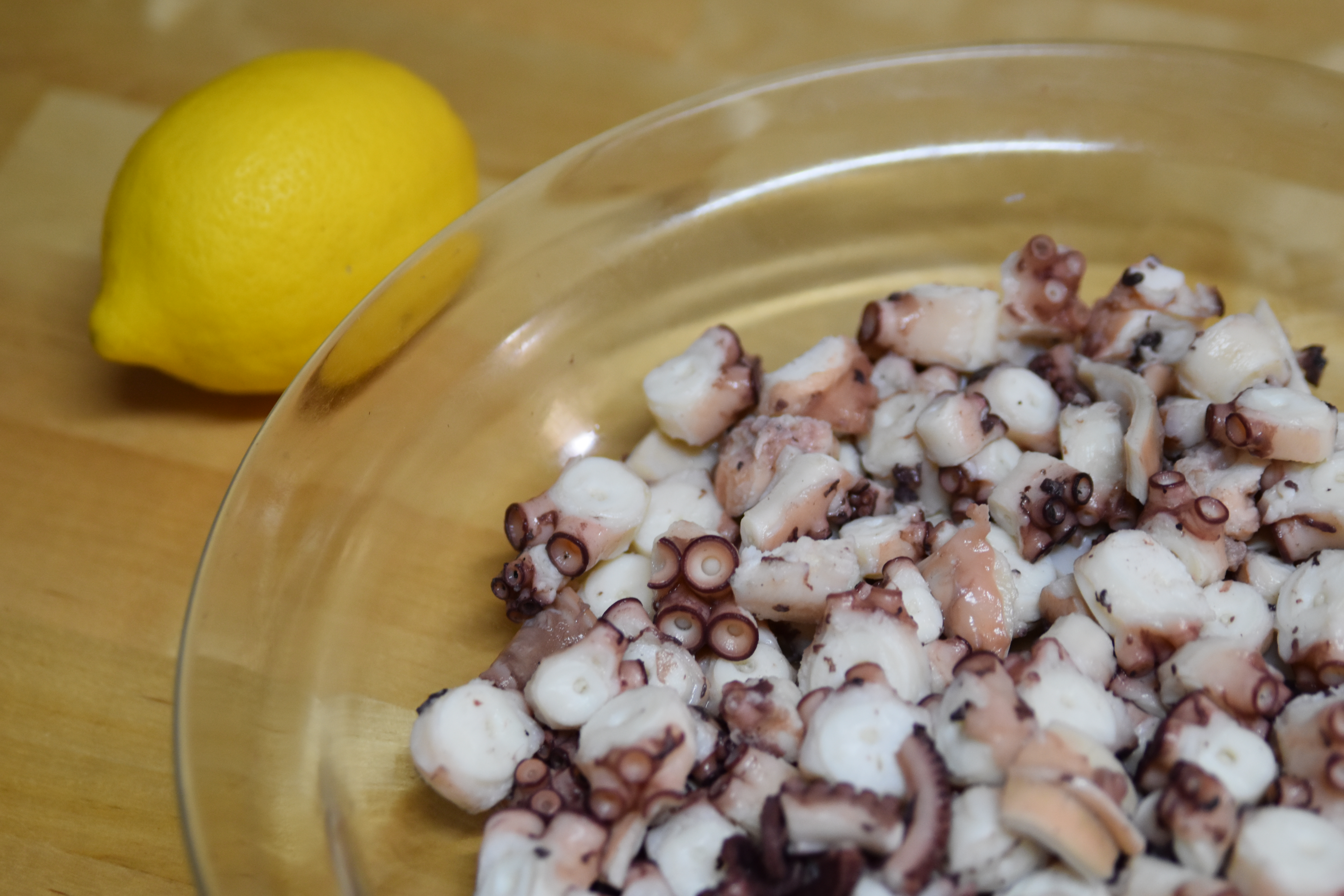 In a glass platter sitting on a wooden table, we find some pieces of white and purple octopus. Next to it, a whole bright lemon is waiting to be squeezed.