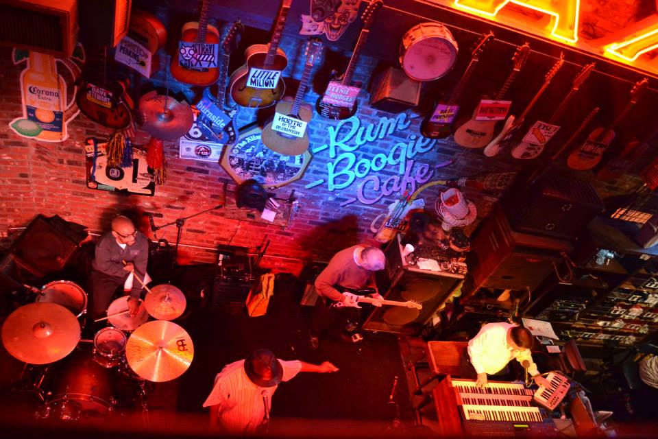 Aerial view of the Rum Boogie Café stage, with four man playing on it: a drummer, a guitarist, a singer with a hat, and a pianist. On the brick wall, the name of the bar is lightened in blue, while the rest of the place is under a red light. Also, about a dozen guitars are hanging from the wall.