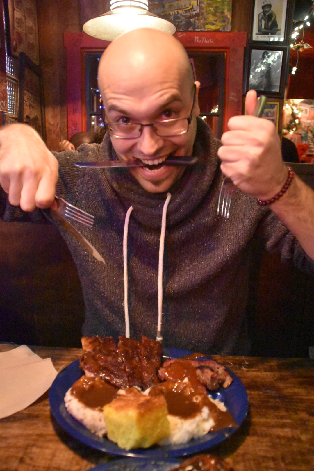 A 30-something year-old man is holding one fork in each hand and a knife between his teeth, smiling in front of a blue dish with mashed potatoes with a brown sauce, a piece of corn bread, half a rack of ribs and some brisket.